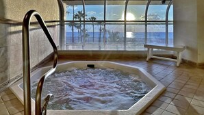 A hot tub with a view of palm trees and the ocean through large windows.