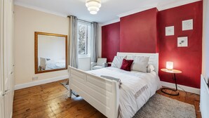 Et soveværelse med en hvid seng, en rød accentvæg, et lille natbord med en lampe og et stort spejl på væggen.