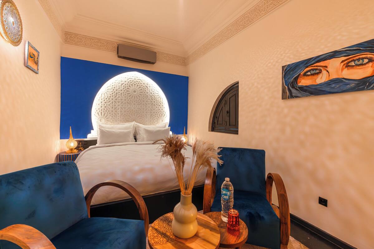 A hotel room with a bed, blue upholstered chairs, a small table with a vase and a bottle, and a large decorative wall piece.