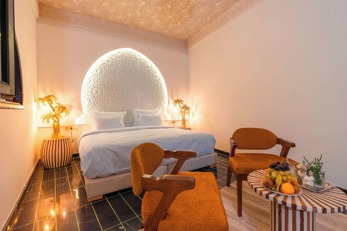 A modern bedroom with a large bed, two nightstands, a round mirror, a wooden chair, and a small table with fruit.
