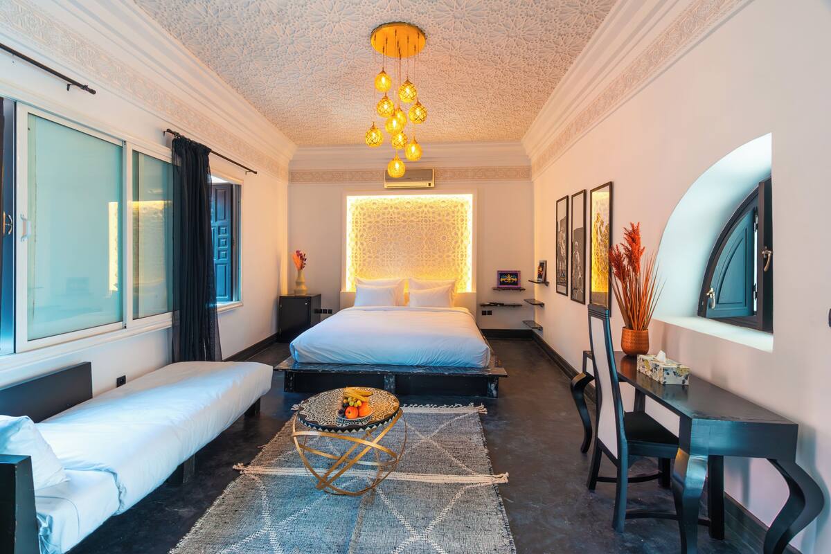A modern hotel room with a large bed, a small round table with fruit, a desk with a chair, and a large window with curtains.