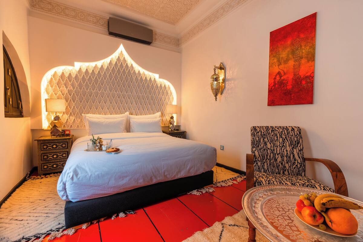 A hotel room with a large bed, a patterned chair, a small table with fruit, and a red floor.