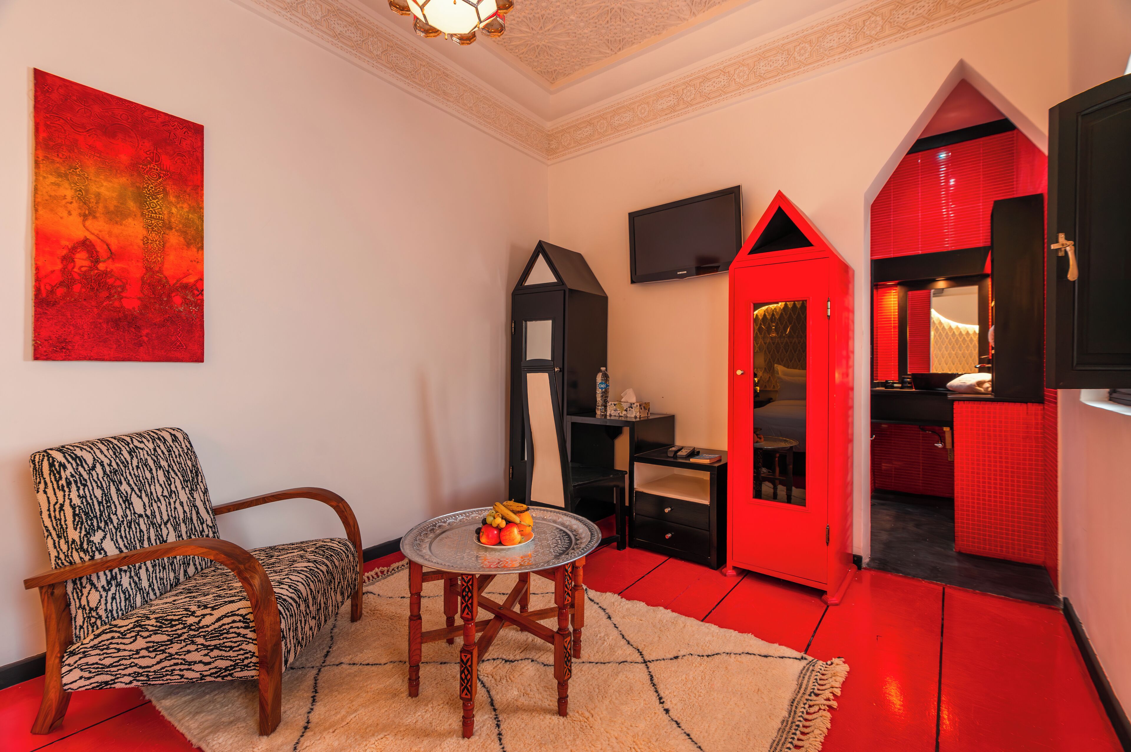 Un espacio habitable compacto con una pared de acento roja, un televisor negro montado en la pared, una mesita redonda con fruta y un sillón con estampado.