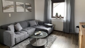 En moderne stue med en grå hjørnesofa, et glasbord og udsigt til det fri gennem et vindue med grå gardiner.