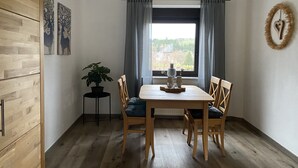Spisested med et træbord og stole, en potteplante og udsigt gennem et vindue med gardiner.