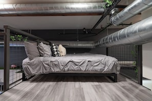 A modern bedroom with a bed, grey bedding, and a view through a window with plants.