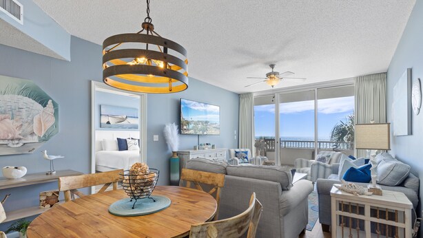 A spacious living area with a wooden dining table, grey sofas, and a large window offering a view of the ocean.