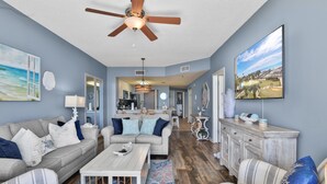 A spacious living room with a ceiling fan, a large TV, and a view of the kitchen in the background.