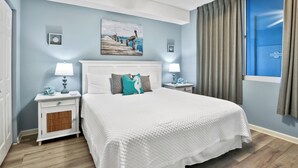 A bedroom with a large bed, white nightstands, a ceiling fan, and a window with curtains.