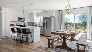 A modern kitchen with white cabinets, a central island with bar stools, a large dining table with benches, and a view of the outdoors through a sliding glass door.