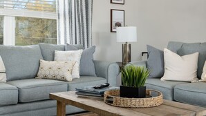 A living room with a grey sofa, a coffee table, and a lamp. There are framed pictures on the wall and a window with blinds.