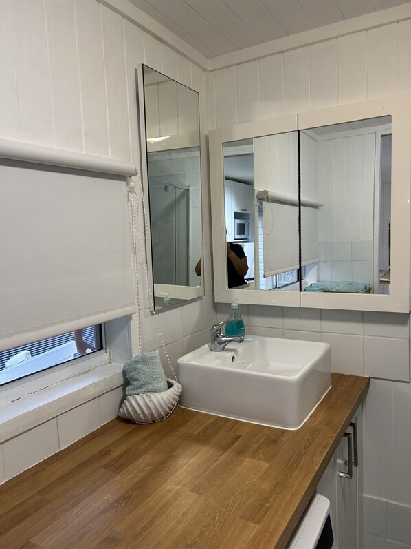 Un baño con tocador de madera, lavabo blanco, espejo y ventana con persianas.