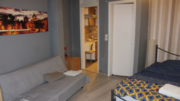 A bedroom with a grey sofa, a bed with a patterned duvet, a painting of a cityscape, and a kitchenette in the background. - Remarkable 1-bed Apartment in Thessaloniki (Thessaloniki)