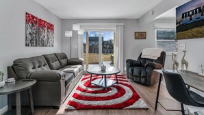 A modern living room with a grey sofa, a black leather chair, a round coffee table, and a red and white rug. There is a painting on the wall and a window with a view of the outdoors. - Fenton Flats- Suite 1 With On Site Hot Tub and Personal Garage (Fenton Township)