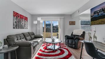 A modern living room with a grey sofa, a black leather chair, a round coffee table, and a red and white rug. There is a painting on the wall and a window with a view of the outdoors.