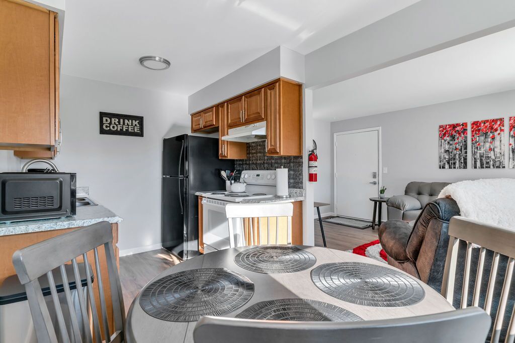 Une cuisine moderne avec des meubles en bois, un réfrigérateur noir et un four à micro-ondes. On y trouve une table à manger avec des sets de table et une chaise. Un panneau sur le mur porte l’inscription : « BUVEZ DU CAFÉ ».