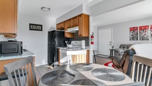 A modern kitchen with wooden cabinets, a black refrigerator, and a microwave. There's a dining table with placemats and a chair. A sign on the wall reads "DRINK COFFEE." - Fenton Flats- Suite 1 With On Site Hot Tub and Personal Garage (Fenton Township)