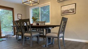 Un coin repas avec une table en bois et quatre chaises, une fenêtre donnant sur les arbres et un tableau encadré au mur.