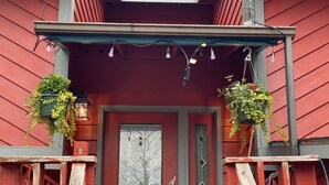 Une maison en bois rouge avec une porte rouge foncé, deux jardinières suspendues et des guirlandes lumineuses au-dessus de la porte.