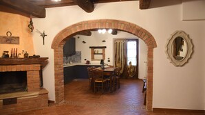 A rustic interior with a brick fireplace, a dining area, and a kitchen visible through an archway.