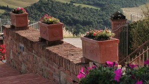 A stone staircase leading to a terrace with terracotta planters containing flowers, overlooking a valley with rolling hills and a distant mountain range.