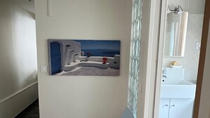 Un pasillo con una silla azul, un cuadro de una escena costera y un baño con lavabo y espejo blancos.
