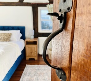 A wooden door with a metal handle, a bed with white bedding, a nightstand with a lamp, and a window with a view of the outdoors. - Log Burner and Beamed Ceilings-king Size Bed (Worcester)