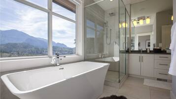 A modern bathroom with a freestanding bathtub, a glass-enclosed shower, and a view of mountains through large windows.