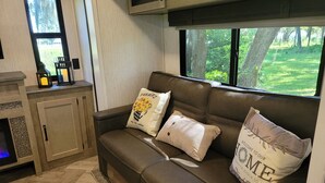 Un rincón acogedor con un sofá de cuero marrón, dos cojines decorativos, un mueble bajo y una ventana con vistas a un paisaje verde.