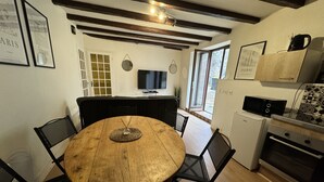 Un coin repas comprenant une table ronde en bois, des chaises noires, un petit objet décoratif posé sur la table, un téléviseur fixé au mur et un espace cuisine équipé d’un four à micro-ondes et d’un four traditionnel.