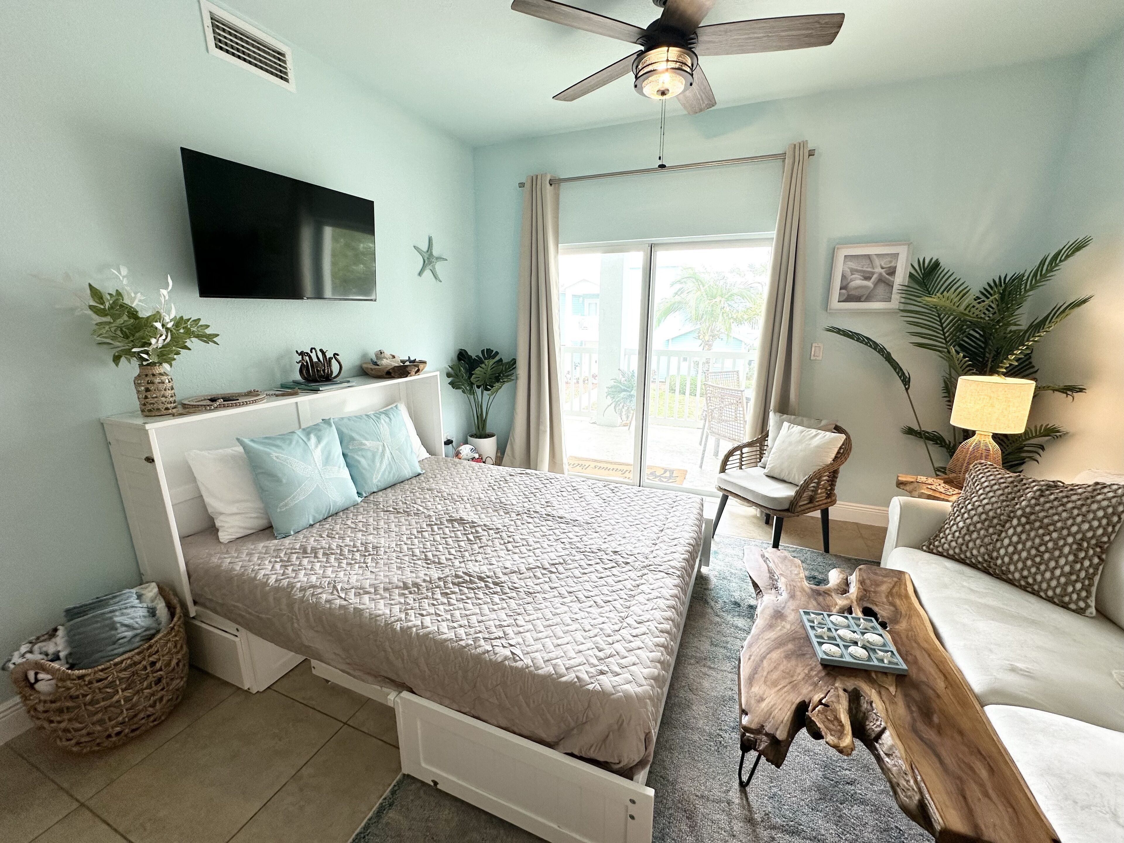 A bedroom with a bed, a ceiling fan, a TV, a window with curtains, a plant, and a wooden coffee table.