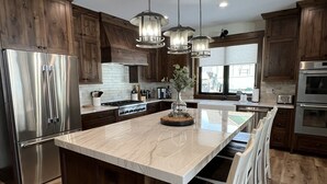 A modern kitchen with wooden cabinets, stainless steel appliances, a marble island, and pendant lighting. - New Luxurious waterfront cabin on Henry's Lake. 26 minutes to West Yellowstone  (Island Park)