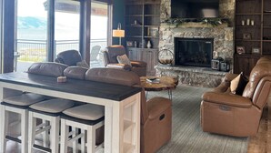 A spacious living room with a stone fireplace, leather seating, a large TV, and a view of the outdoors. - New Luxurious waterfront cabin on Henry's Lake. 26 minutes to West Yellowstone  (Island Park)