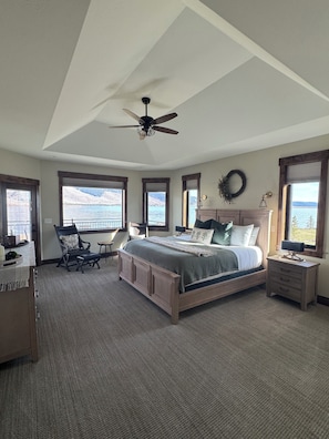 A bedroom with a large bed, a ceiling fan, a desk, and a view of a lake.