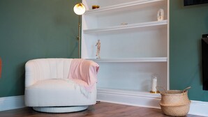 Une pièce avec un fauteuil blanc, un panier en osier et une étagère blanche ornée d’objets décoratifs.