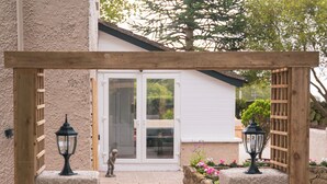 Un chemin mène à un bâtiment blanc doté d’une entrée avec une pergola en bois, de murs en pierre et d’une petite statue.
