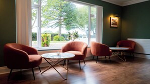 Une pièce avec deux tables rondes et trois fauteuils rouges, de grandes fenêtres donnant sur un paysage verdoyant, et un tableau encadré accroché au mur.
