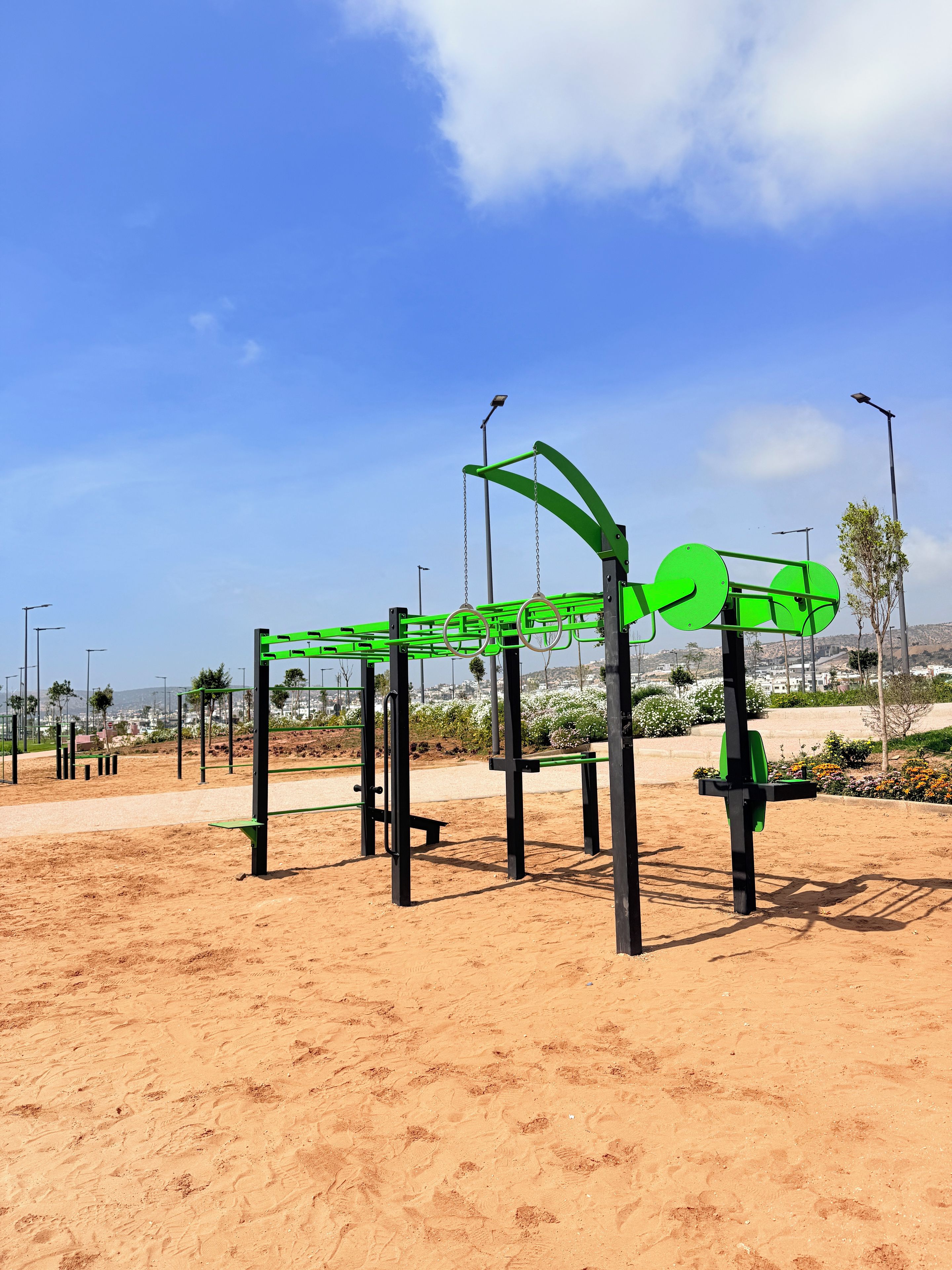 Una estructura para ejercicios al aire libre con marco metálico y barras verdes, sobre un terreno arenoso y un cielo despejado.