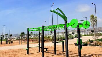 Una estructura para ejercicios al aire libre con marco metálico y barras verdes, sobre un terreno arenoso y un cielo despejado.