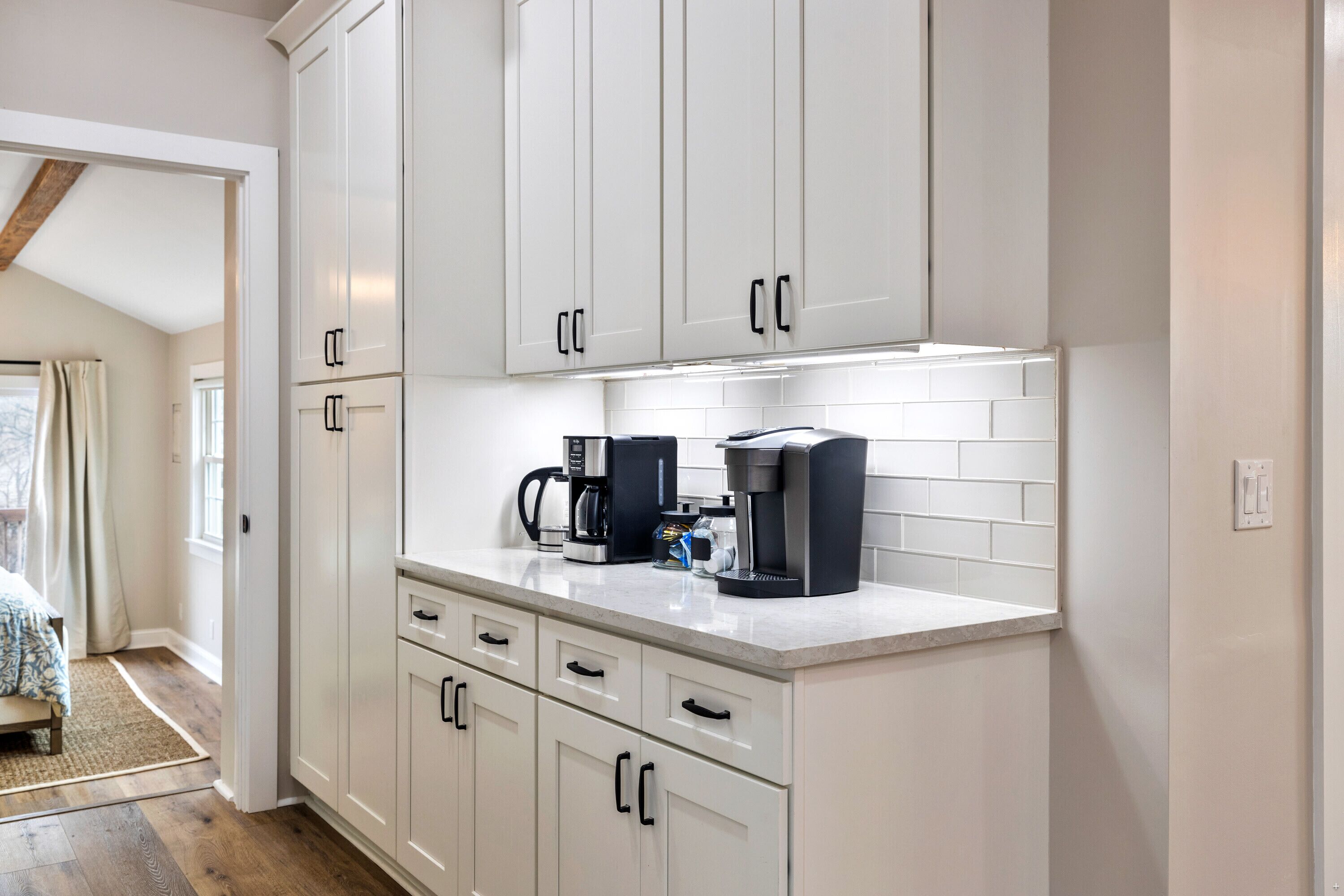 Una cocina moderna con armarios blancos, una encimera con cafetera y vistas a un dormitorio con cama y ventana.