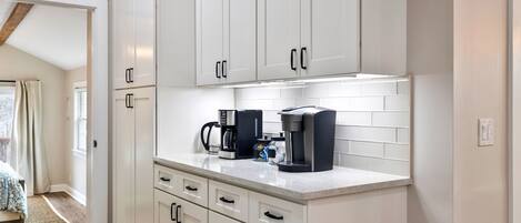 Una cocina moderna con armarios blancos, una encimera con cafetera y vistas a un dormitorio con cama y ventana.