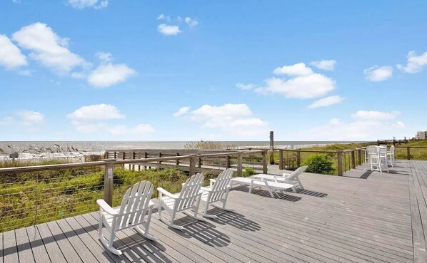 A wooden deck with white Adirondack chairs overlooking a beach.