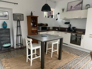 A compact kitchen with white cabinets, a dining table with chairs, a standing fan, and a microwave. - Maison Jardin RDC Proche Plages et Commerces (Trébeurden)