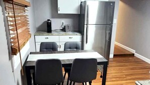 A compact kitchen with a refrigerator, a coffee maker, and a small countertop. There are two chairs and a table in the dining area. - Cozy second floor unit in the heart of Downtown Charlevoix, 1BR / 1BA sleeps 4 
 (Charlevoix)