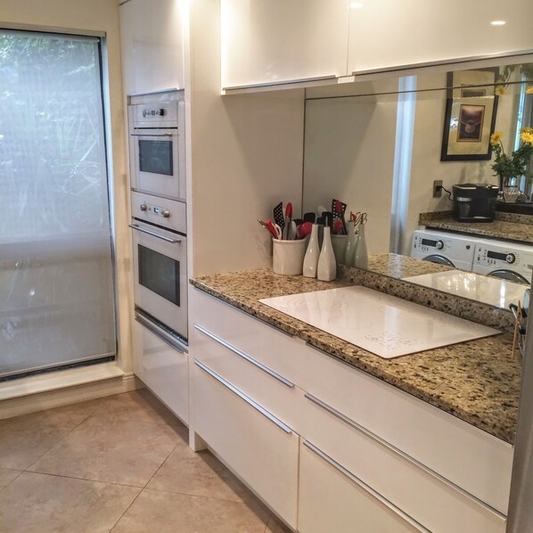Una cocina con encimera de granito, gabinetes blancos, horno empotrado, un lavabo y un espejo grande sobre la encimera.