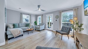 A modern living room with a grey sofa, a grey armchair, a wooden coffee table, and a ceiling fan. There is a large window with curtains, a door with a glass panel, and a wall-mounted light fixture. - Highly Rated Cozy 30A Blue Mountain Beach Escape! (Santa Rosa Beach)