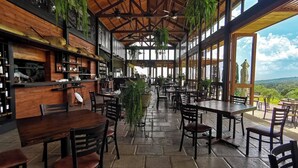An indoor dining area with wooden tables and chairs, a bar area with shelves of bottles, and large windows offering a view of the outdoors. - Finca San Cayetano (Alotenango)
