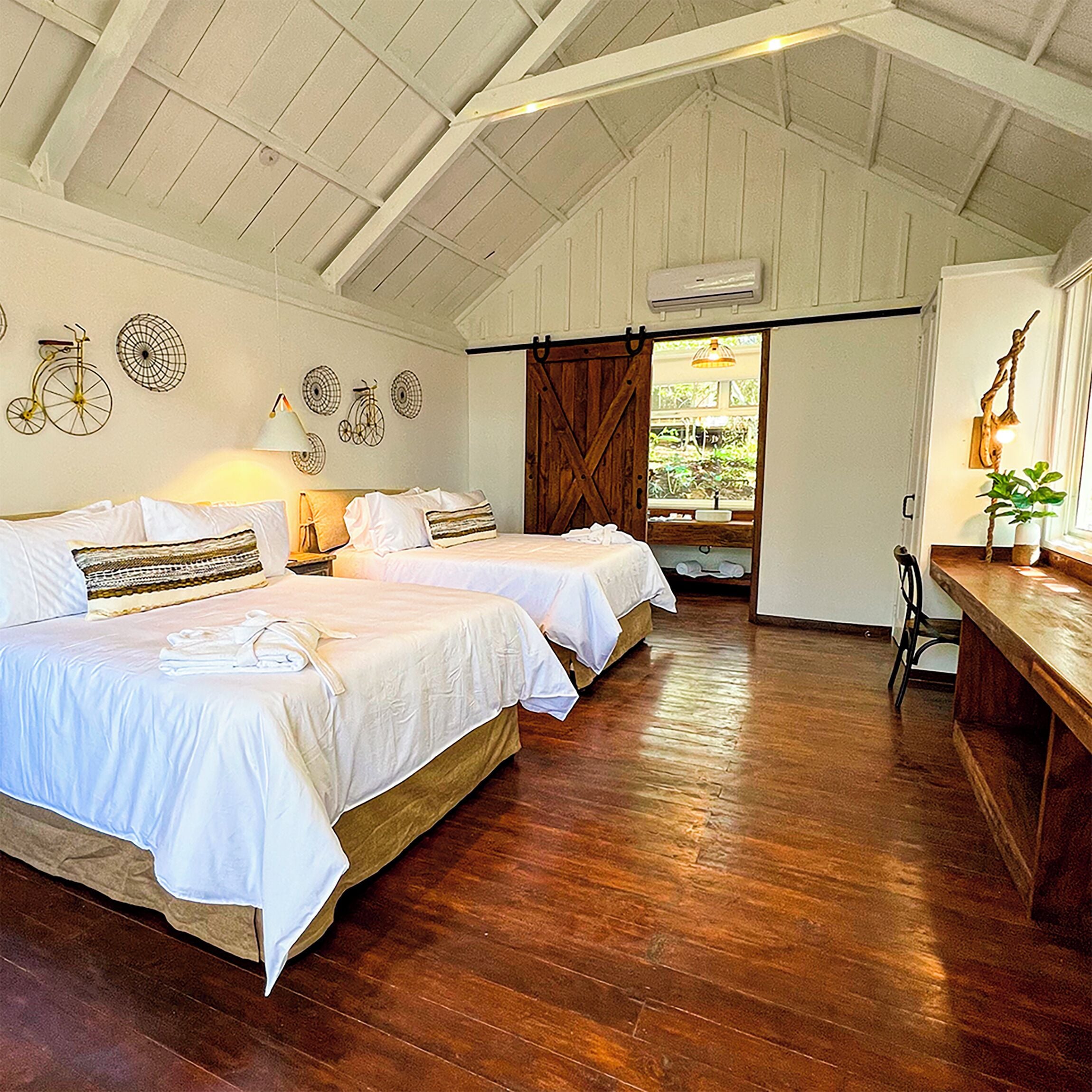 A room with two beds, wooden flooring, a desk, and a large window.
