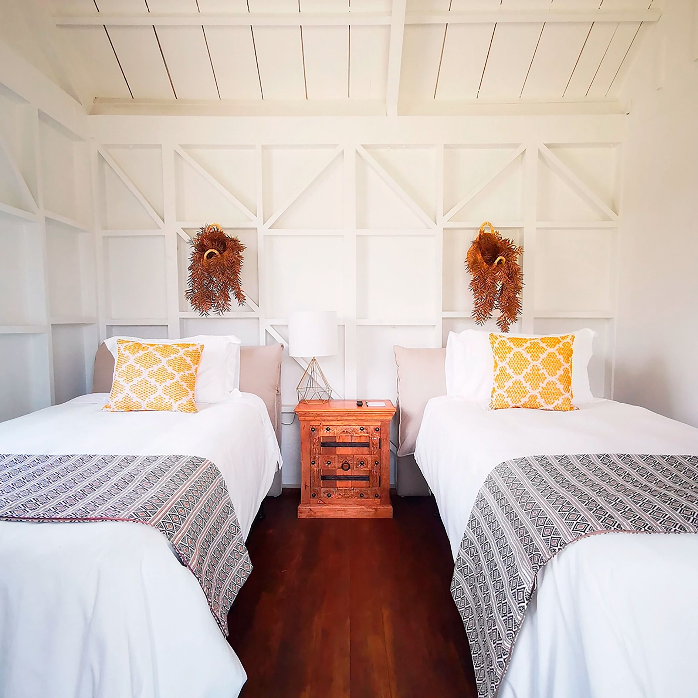 Habitación de hotel con dos camas, ropa de cama blanca, almohadones estampados, una mesita de noche de madera y una lámpara fijada a la pared.