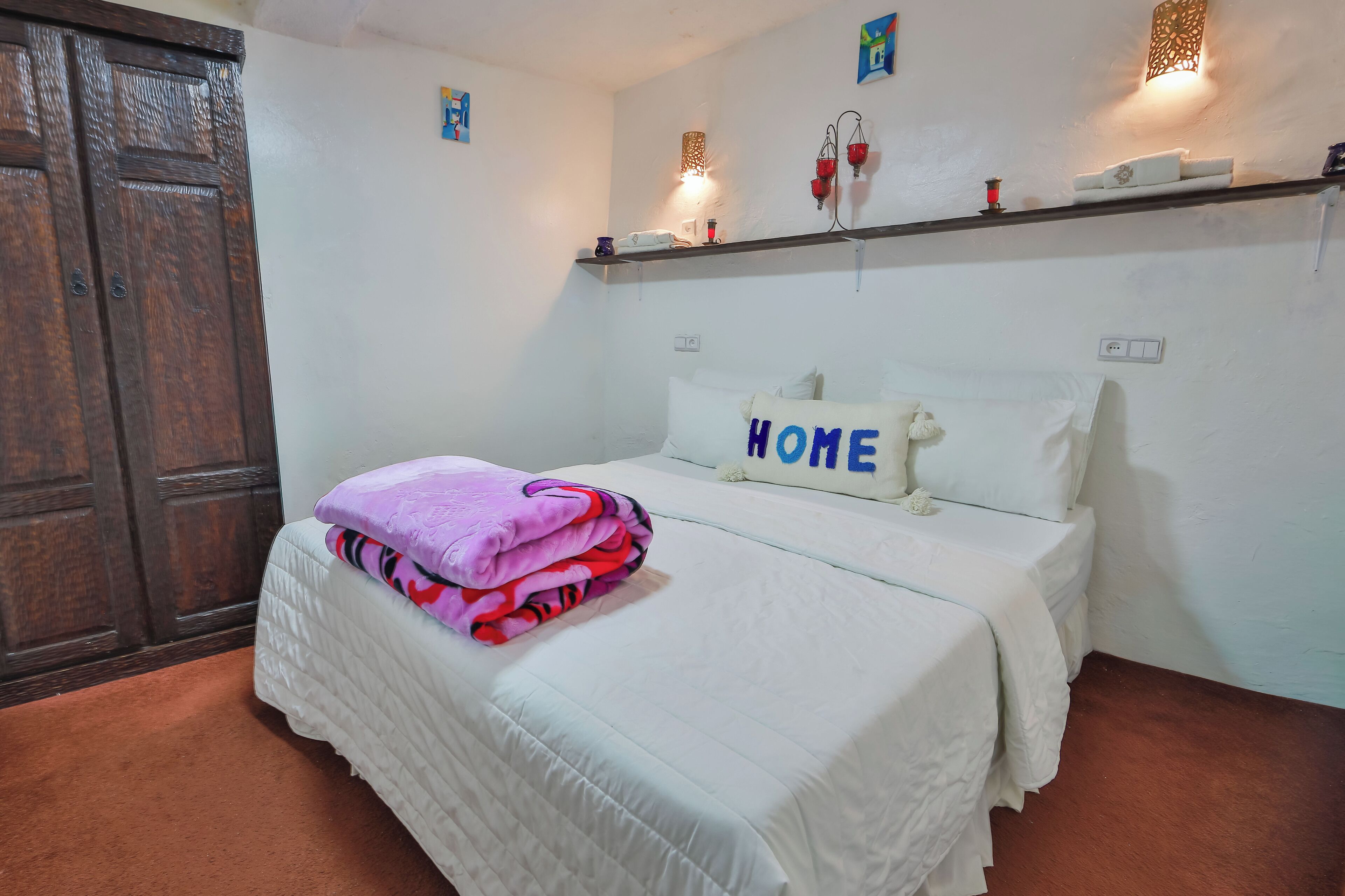 A bedroom with a bed, a wardrobe, a shelf with decorative items, and a wall-mounted light fixture.
